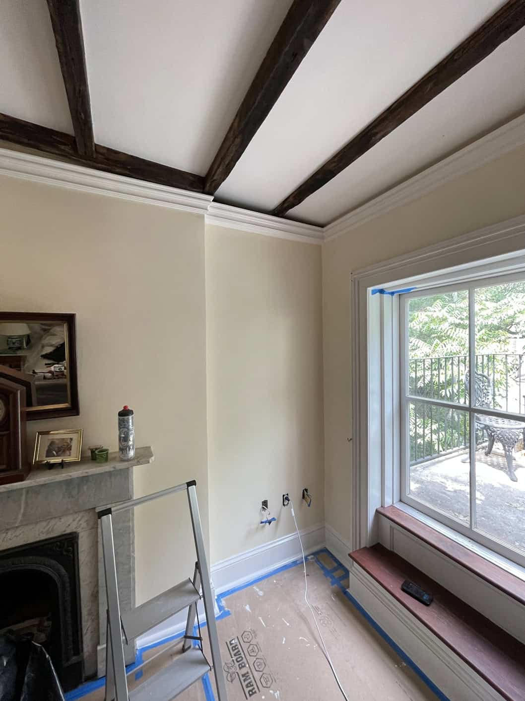 A cozy corner with a fireplace and window seat is prepped for a makeover by painting contractors NYC. A small ladder, painter's tape on the walls, and protective covering on the floor set the scene, while exposed beams add charm. Through the window, trees paint a serene picture outside.