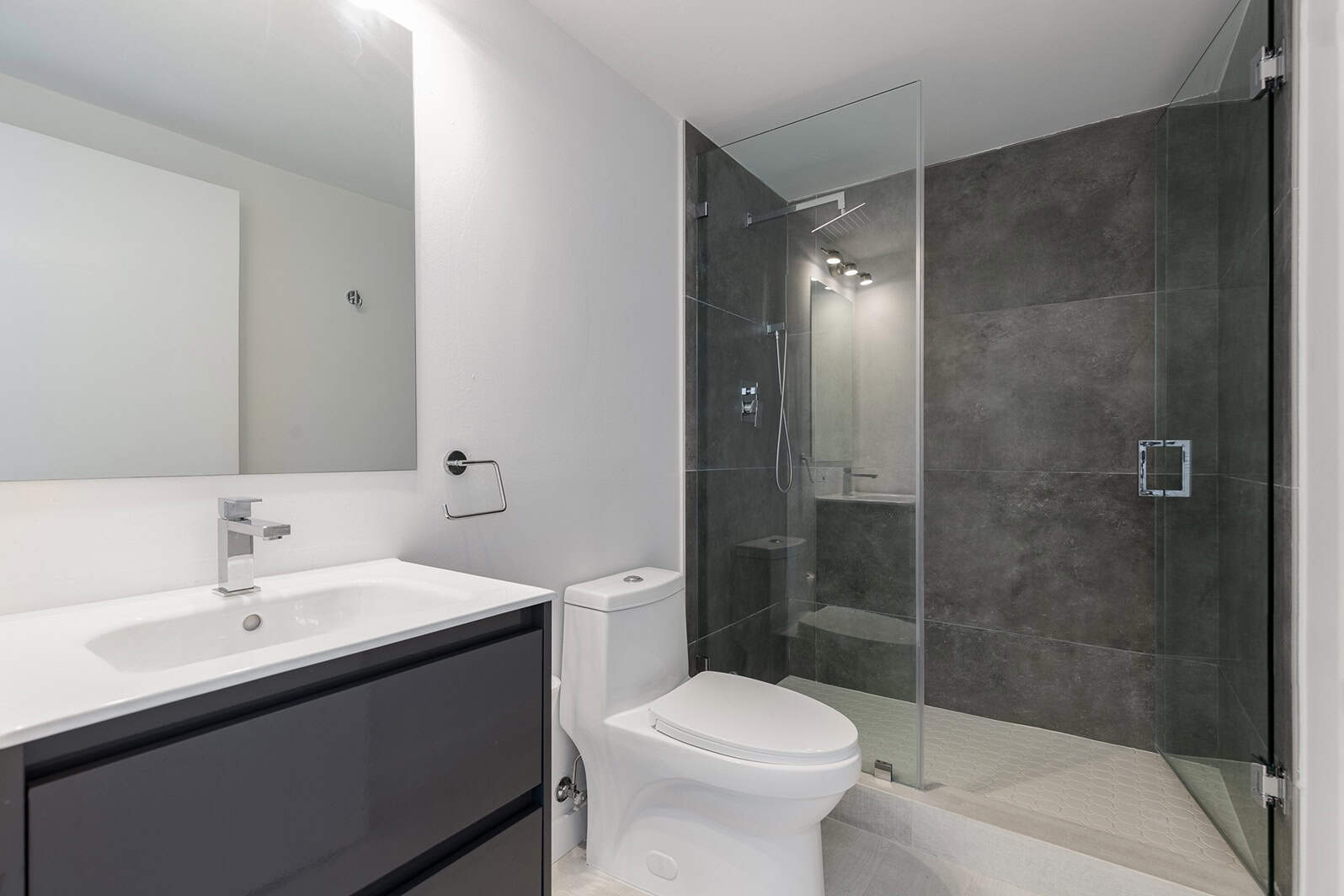 Modern bathroom with a white sink and dark vanity on the left, a toilet in the center, and a glass-enclosed shower with dark tiles on the right. Walls are crisp white, flooring is light, and bright minimalist lighting offers an elegant touch. Perfect for those seeking painting services NYC to refresh their space.