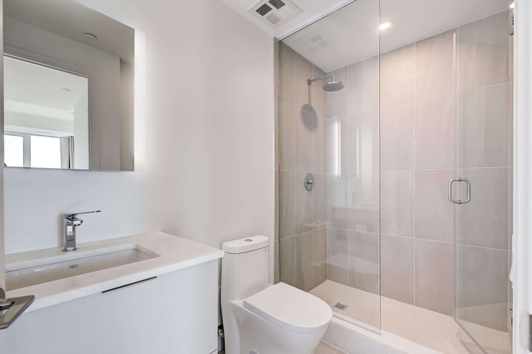 Modern bathroom with a glass-enclosed shower, beige tiles, and a white sink with a silver faucet. A wall-mounted mirror hangs above, beside the toilet. Expertly lit through a small window, this sleek design might even inspire you to consider painting services NYC for your next project.