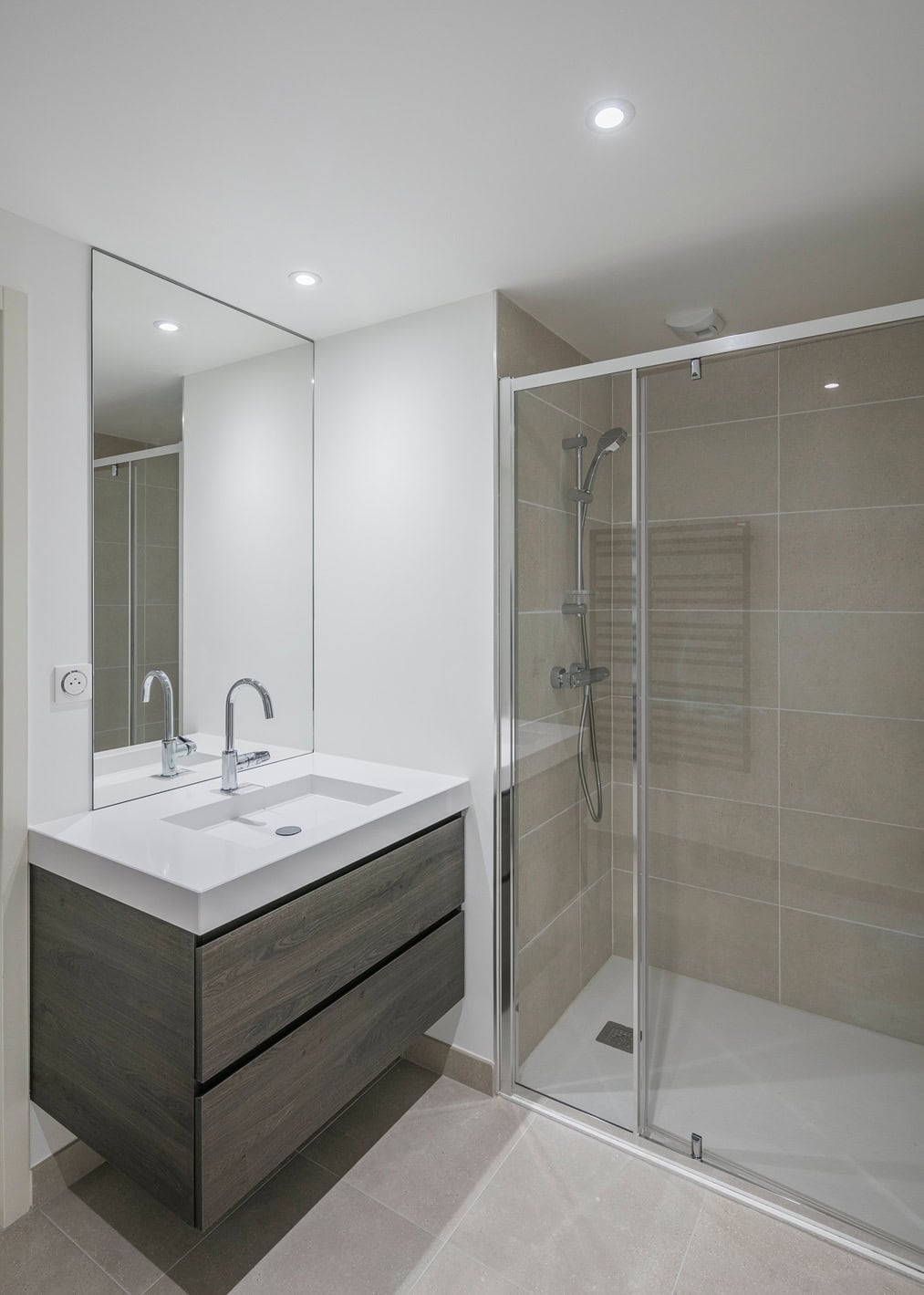 Minimalist bathroom with a large mirror above a dark wood double-sink vanity and spacious glass shower. The walls and floor, expertly tiled in light gray by painting contractors NYC, are illuminated by recessed lighting, creating an elegant ambiance.