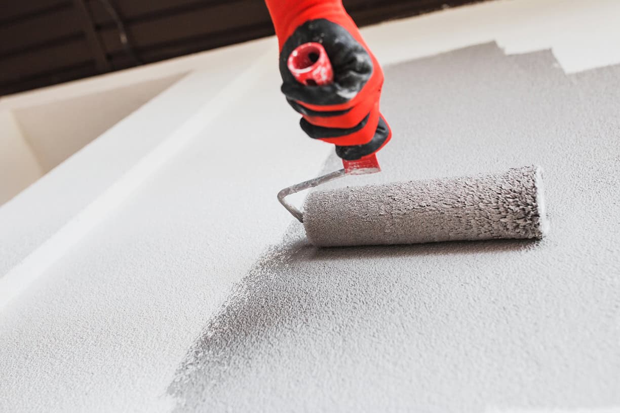 A person wearing red gloves uses a paint roller to apply light gray paint on a wall, reminiscent of the expert finish painting contractors NYC provide. The roller is partially covered with wet paint, revealing a clear distinction between painted and unpainted areas.