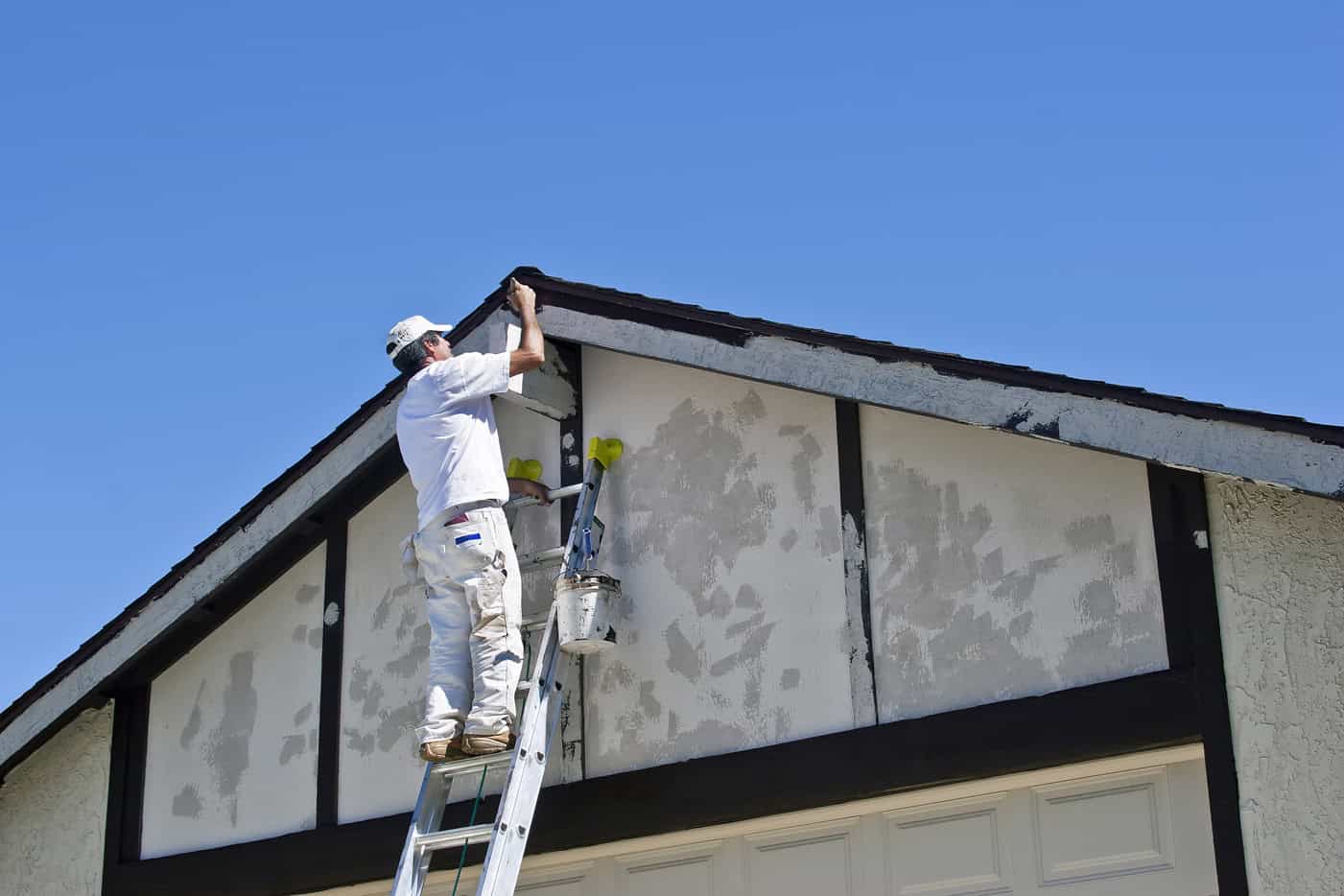 A person in white work clothes stands on a ladder, painting the eaves of a house under a clear blue sky. They hold a small paintbrush, reaching towards the roofline. A paint bucket hangs from the ladder as they showcase expert skills akin to top-notch painting contractors NYC offers.