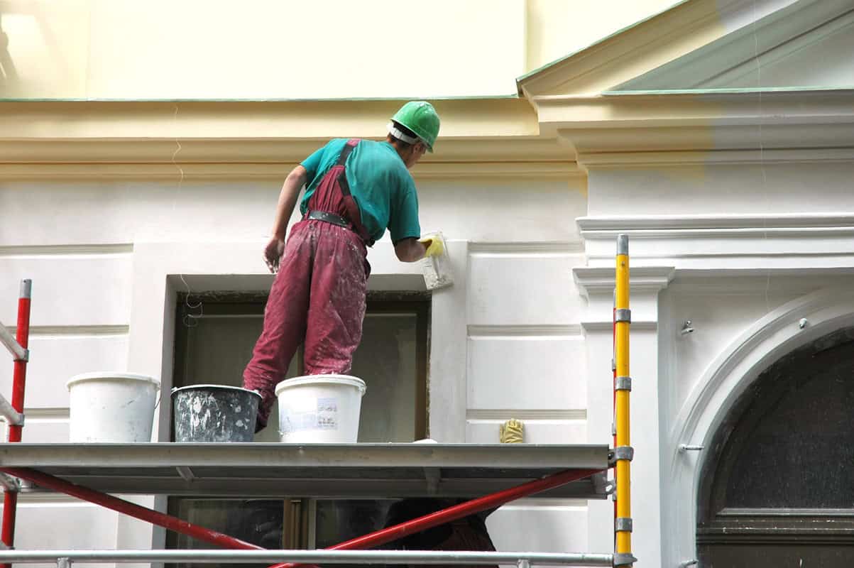 A construction worker in a green hard hat and red overalls stands on scaffolding while painting the exterior of a building, showcasing top-notch painting services NYC. He uses a roller brush on a light-colored wall with a window below. Buckets are placed on the scaffolding.