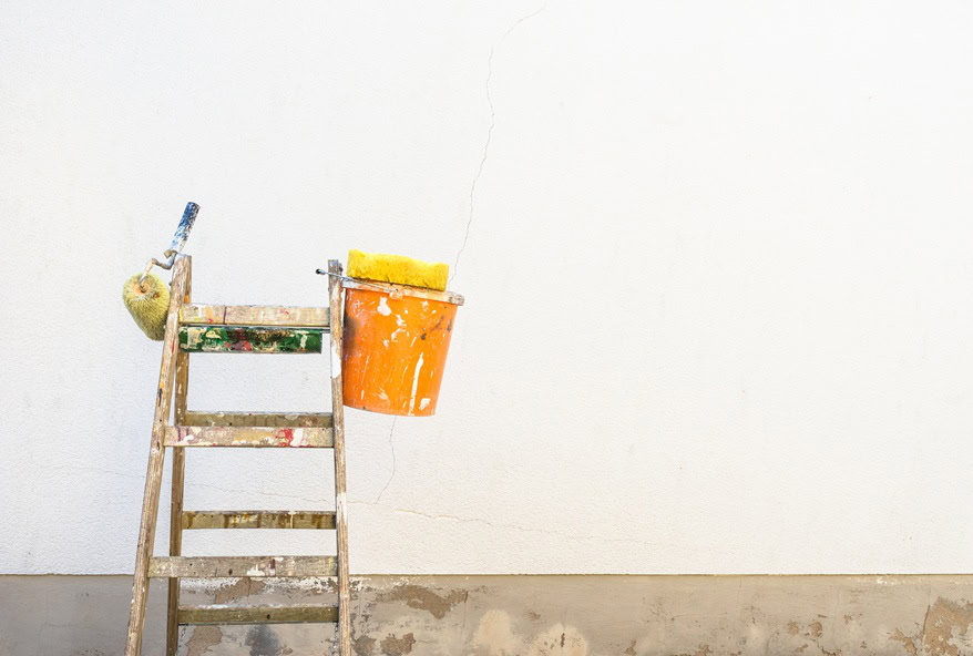 A wooden ladder stands against a white wall with visible cracks, hinting at the need for painting services NYC. On the ladder, there's an orange bucket and a paint roller. The bottom of the wall shows patches of exposed, unfinished surface.