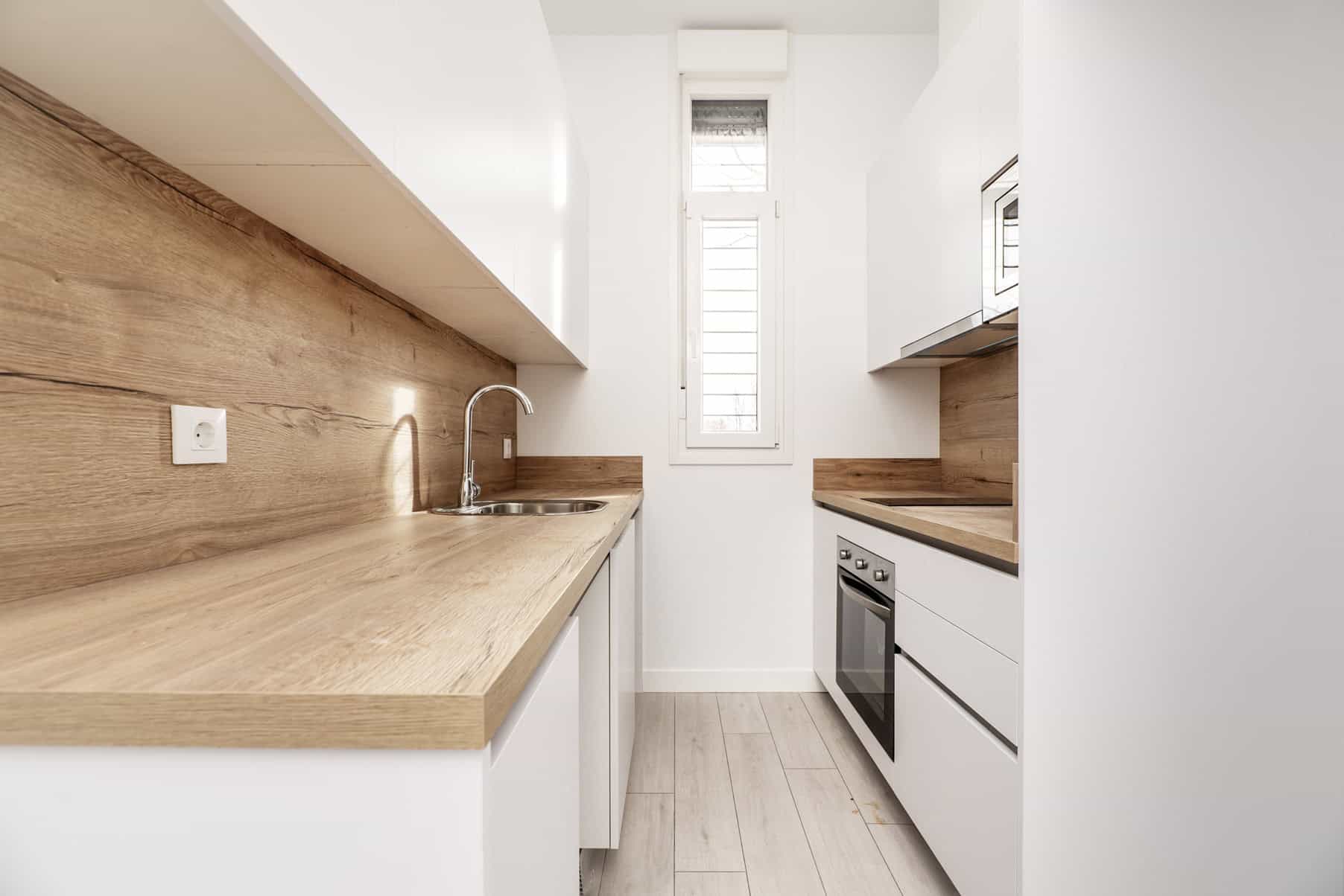 Minimalist kitchen with white cabinets, light wood countertops, and a wood backsplash. A stainless steel sink and stove are visible. Natural light streams in from a tall, narrow window, illuminating the bright space—perfect for showcasing work by top painting contractors NYC.