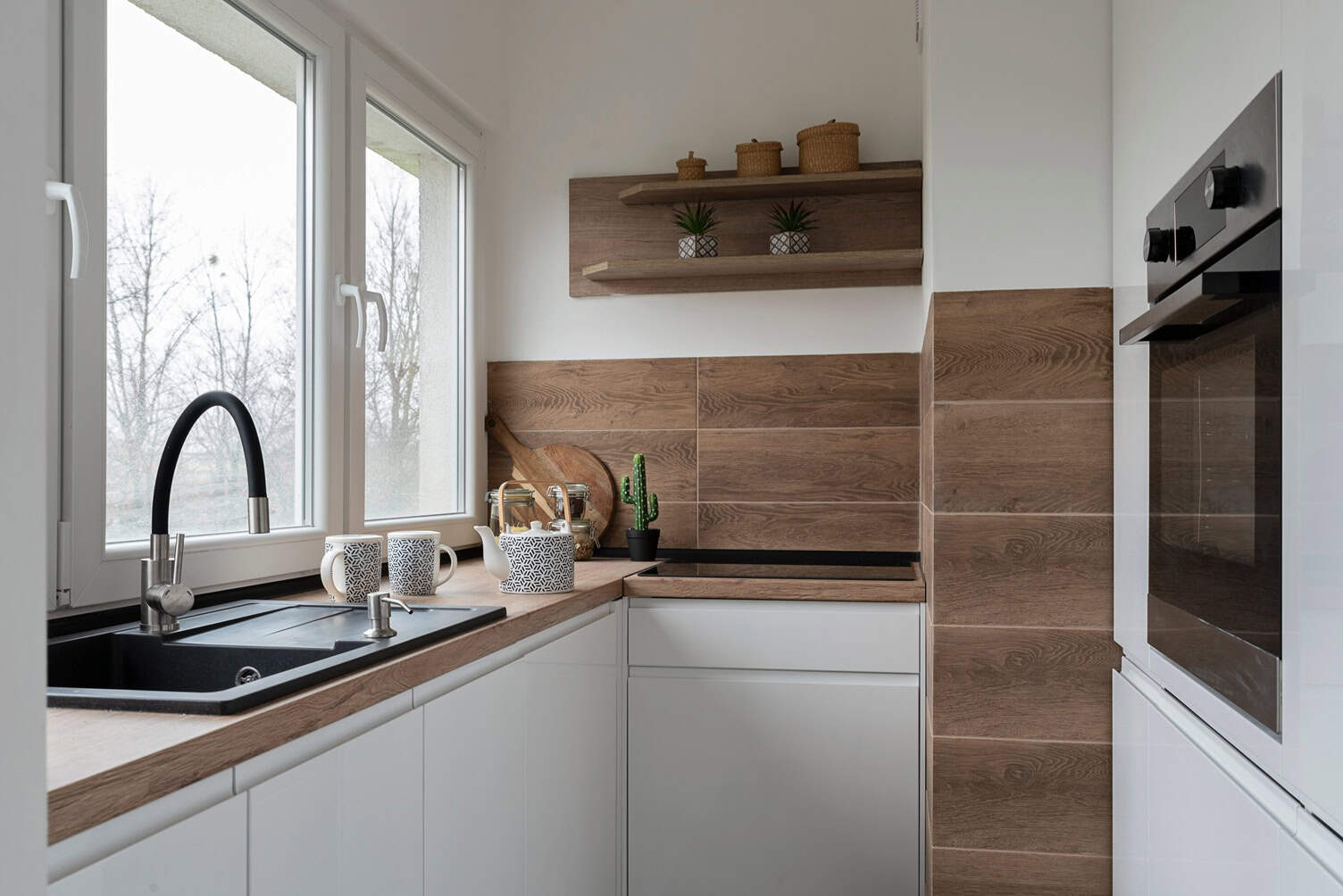Modern kitchen with white cabinets and wood accents. Features a black sink, oven, stovetop, and natural light from large windows. Open shelves hold small pots and decor. Minimalist design with clean lines and earthy tones achieved by top painting contractors NYC.