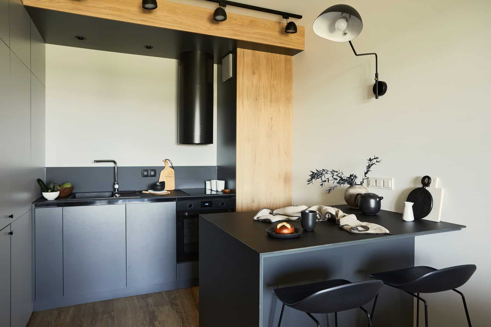 Modern kitchen with dark cabinetry and wooden accents. A black oven and sleek exhaust hood are set into the walls. Featuring a breakfast bar with black stools, crockery, and fabric. Minimalistic decor includes a potted plant on the counter, expertly painted by top painting contractors in NYC.
