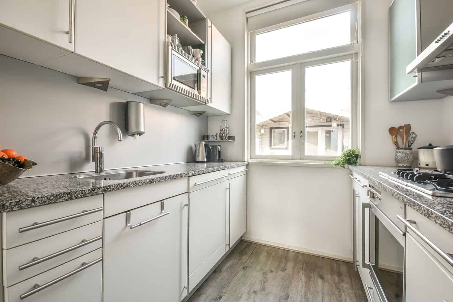 A modern kitchen with white cabinets and granite countertops basks in sunlight streaming through a large window. A fruit bowl sits on the counter, surrounded by utensils and appliances. The wood-finish floor gleams, thanks to painting contractors NYC who ensure every detail is perfect.