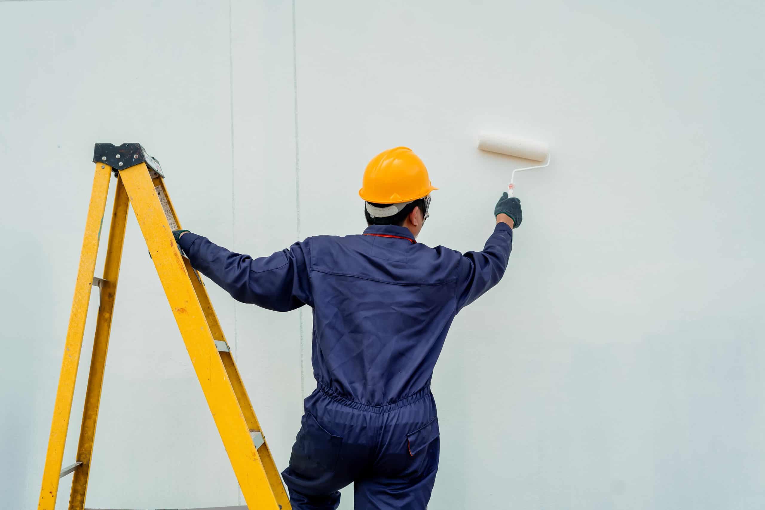 A person in a yellow hard hat and blue coveralls expertly uses a paint roller on a white wall, standing beside a yellow ladder. They represent the top painting contractors NYC relies on for quality work.