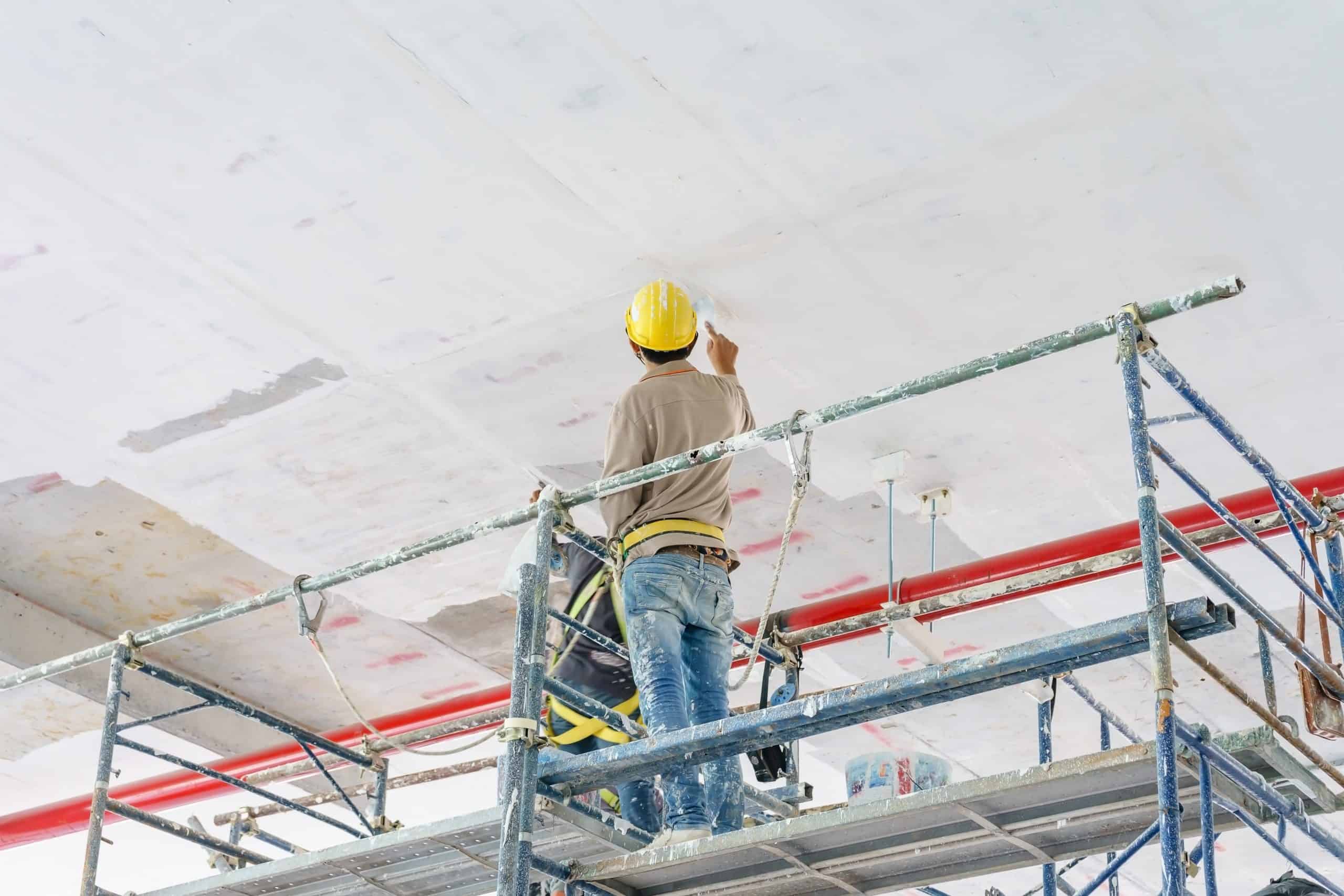 A construction worker wearing a yellow hard hat stands on scaffolding, painting a large white ceiling. Red pipes loom above him, and paint supplies are scattered nearby. Dressed in a brown shirt and blue jeans, he exemplifies the expertise of painting contractors NYC relies on.