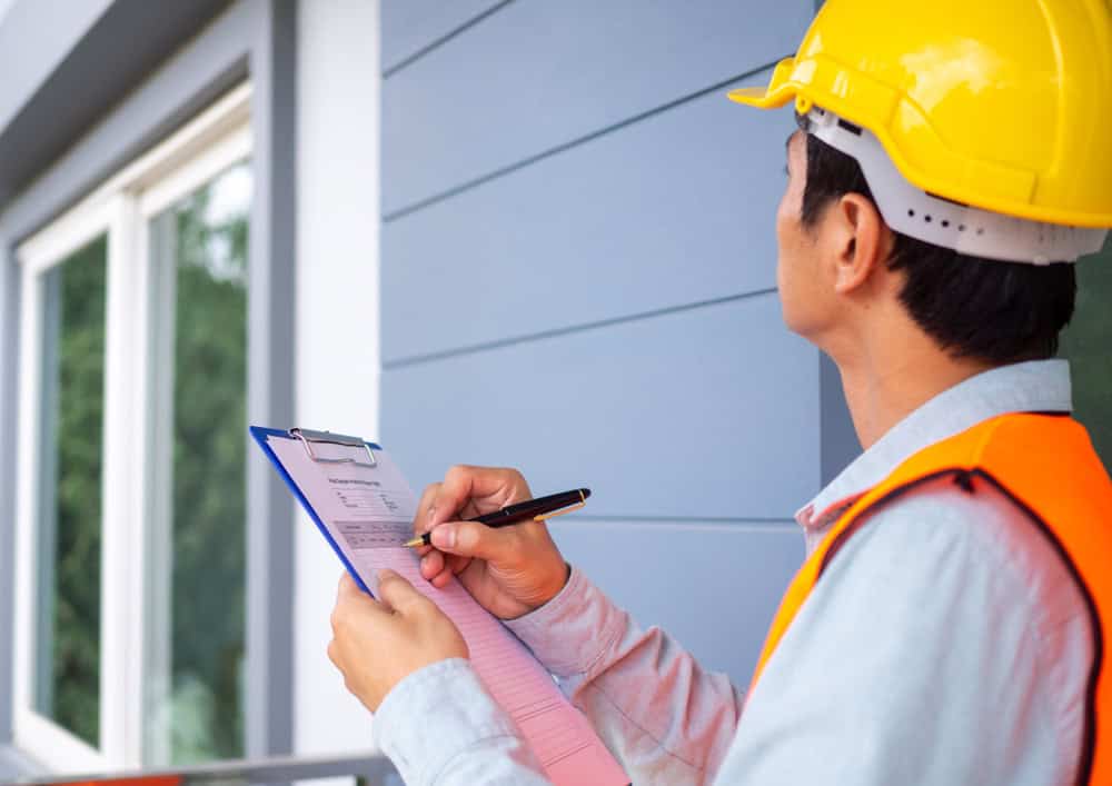 A person wearing a yellow hard hat and orange safety vest is holding a clipboard and pen, facing a modern building with horizontal paneling and a window. The person appears to be inspecting or taking notes.