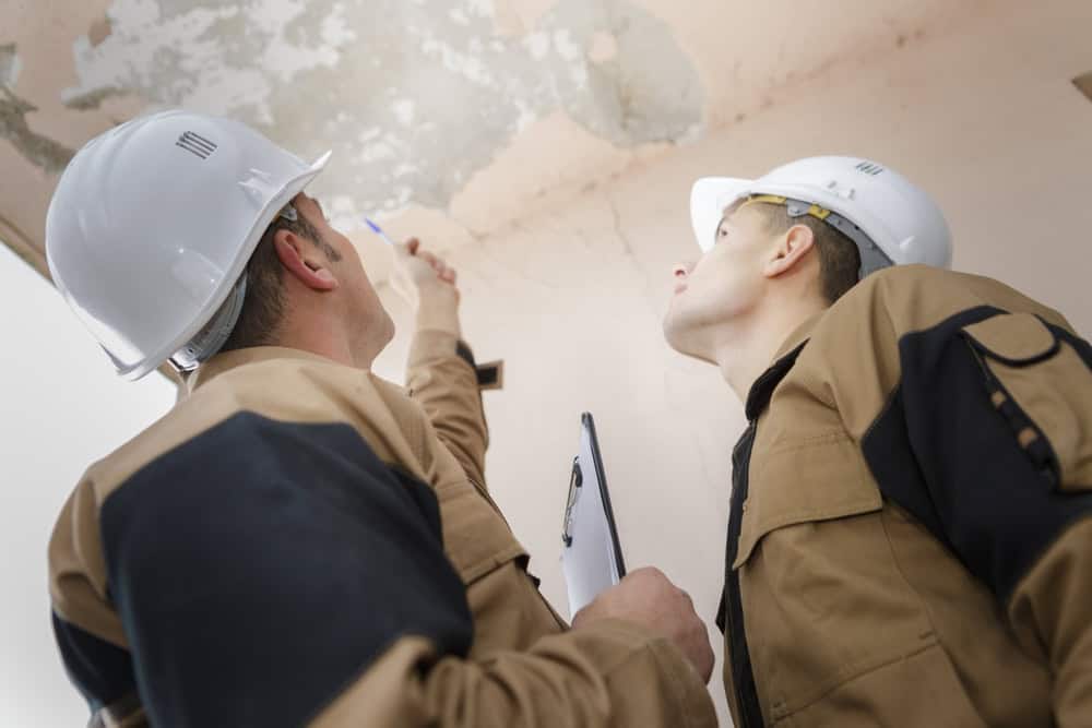 Two construction workers wearing helmets and brown uniforms inspect a ceiling. One points upwards with a pen, while the other holds a clipboard. They appear to be assessing damage or wear.