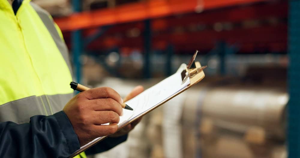 A person in a high-visibility jacket writes on a clipboard with a pen. The background is a blurred view of shelves in a warehouse.